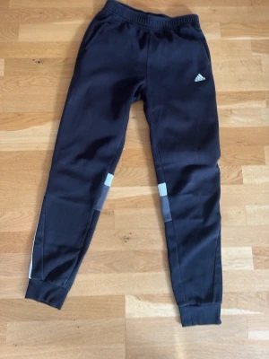 Svarta mjukisbyxor från Adidas - Säljer ett par svarta mjukisbyxor från Adidas med smal passform och vita detaljer vid knäna. Byxorna har resår i midjan och muddar vid bensluten. Perfekta för gymmet eller att vila i hemma.