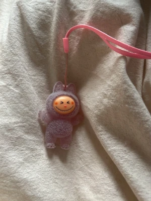Lila fluffig figur-nyckelring - Söt nyckelring med en liten figur i lila fluffigt material och ett glatt ansikte. Figuren har kattöron och en rosa handledsrem, perfekt att hänga på väskan eller nycklarna. Materialet är mjukt och plyschliknande.