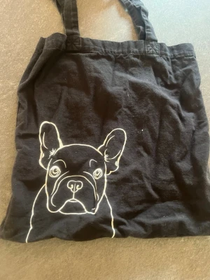 Svart tygkasse med hundmotiv - Säljer en svart shoppingväska i tyg med ett vitt tryck av en fransk bulldogg på framsidan. Väskan har två handtag och är perfekt för att bära böcker, mat eller annat du behöver. Enkel och snygg design som sticker ut.