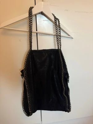 Svart handväska med kedjedetaljer - Cool svart handväska med glansig finish och kraftiga silverfärgade kedjor som axelband och längs kanterna. Väskan har en mjuk, slouchy form och vinrött innerfoder med innerfack. Perfekt för dig som gillar edgy detaljer och rymliga väskor.