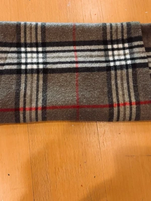 Burberry   - Snygg halsduk i brunt med klassiskt rutmönster i svart, vitt och rött. Materialet känns mjukt och värmande, perfekt för kyliga dagar. Tidlös design som passar till de flesta jackor och stilar. Avslutas med fransar i kanterna.