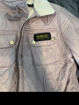 Ljuslila quiltad jacka från Barbour - Quiltad jacka från Barbour i ljuslila med bälte i midjan, fyra fickor framtill och dragkedja. Jackan har en hög krage med vit insida och detaljerade knappar. Perfekt för dig som vill ha en snygg och praktisk jacka.
