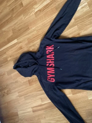 Svart Gymshark hoodie med röd text - Snygg svart hoodie från Gymshark med stor röd logga på bröstet. Klassisk huva med väldigt skönt material och långa ärmar. Perfekt för gymmet eller chill hemma. Tillverkad i mjukt material för maximal komfort.