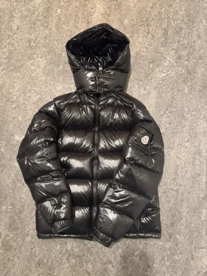 Moncler Maya Jacka - Snygg svart dunjacka från Moncler med glansig finish och huva. Jackan har en klassisk puffer-modell, dragkedja framtill och Moncler-logga på ärmen. Perfekt för kalla vinterdagar och riktigt trendig stil. Skick: 9/10. Mer info i DM!