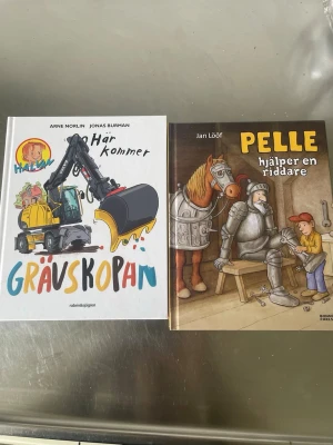 Två barnböcker: Grävmaskin & Riddare - Säljer två färgglada barnböcker: 'Här kommer grävmaskinen' med illustrationer av en stor gul grävmaskin och 'Pelle hjälper en riddare' där Pelle möter en riddare och en häst. Båda böckerna har hårda pärmar och fantasifulla illustrationer.                     Oöppnade 
