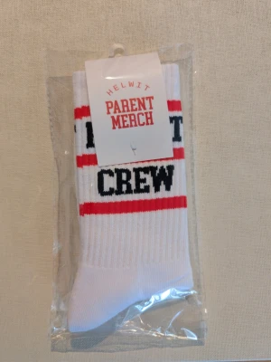Vita strumpor HELWIT Parent Merch - Vita strumpor från HELWIT Parent Merch med texten 'CREW' i svart och röda ränder. Klassisk crew-modell i ribbad bomull som passar perfekt till sneakers eller vardagslooken. Snygg och enkel design med tydlig textdetalj.