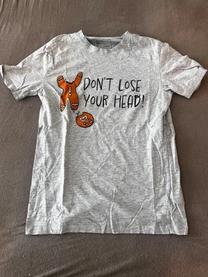 Grå t-shirt med pepparkakstryck - Snygg grå t-shirt med tryck av en pepparkaksgubbe utan huvud och texten 'Don't lose your head!'. Perfekt för dig som gillar humoristiska motiv. T-shirten har rund hals och korta ärmar i mjuk bomull.
