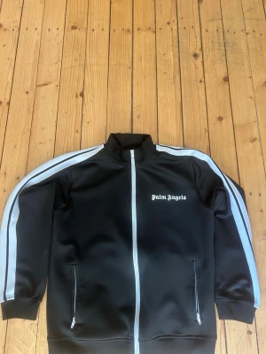 Palm angels - Säljer en svart track jacket från Palm Angels med vita ränder längs ärmarna och vit dragkedja. Jackan har hög krage, två fickor med dragkedja och logga på bröstet. Perfekt för en sportig och trendig look.