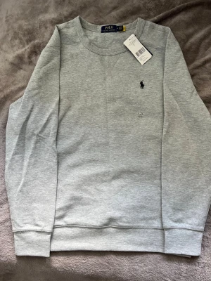Grå sweatshirt från Polo Ralph Lauren - Snygg grå sweatshirt från Polo Ralph Lauren med klassisk rund halsringning och broderad logga på bröstet. Tröjan har ribbade muddar vid ärmslut och nederkant. Perfekt för en avslappnad och stilren look. Aldrig använt den!