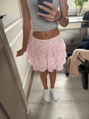 Rosa volangkjol med blommigt mönster - Strechig material så passar även xs använd 1 gång Supersöt kort kjol i ljusrosa med små blommor och flera volanger. Kjolen har en elastisk midja och är perfekt för vår och sommar. Lätt och luftig, passar till både sneakers och sandaler. 