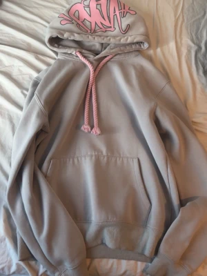 Grå Syna world hoddie - Jag säljer en grå Syna world hoodie som är knappast använd som har storlek S som är svår att hitta nu för tiden, med rosa snören och graffiti-inspirerat tryck i rosa på huvan. Hoodien har en stor magficka och ribbade muddar vid ärmar och nederkant. Perfekt för dig som vill sticka ut med din stil och få massa komplimanger. 