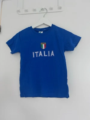 Blå t-shirt med Italia-tryck - Blå t-shirt med korta ärmar och tryck av texten 'ITALIA' samt Italiens flagga på bröstet. Klassisk rund halsringning och enkel design, perfekt för dig som gillar sportig stil eller vill visa ditt stöd för Italien.