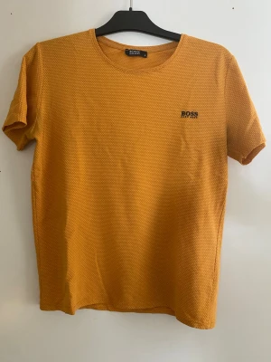 Gul t-shirt från Hugo Boss - Snygg gul t-shirt från Hugo Boss med diskret strukturmönster och liten logga på bröstet. T-shirten har rund halsringning och korta ärmar. Perfekt för dig som vill ha en stilren men ändå unik look. Har ett hål under ena ärmen som lätt kan åtgärdas