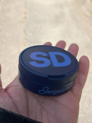 Blå snusdosa med SD-logga - Mörkblå snusdosa i plast med stor SD-logga i ljusblått på locket och texten 'Sverigedemokraterna' på sidan. Perfekt för dig som vill ha en unik accessoar med politisk touch. Praktisk och enkel att ta med sig i fickan.