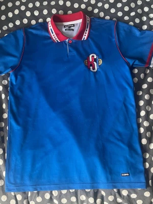 Blå pikétröja från COPA - Blå pikétröja med röd och vit krage, röda detaljer längs ärmarna och broderat H-märke på bröstet. Tillverkad i ett sportigt material och har korta ärmar. Perfekt för dig som gillar sportig stil.
