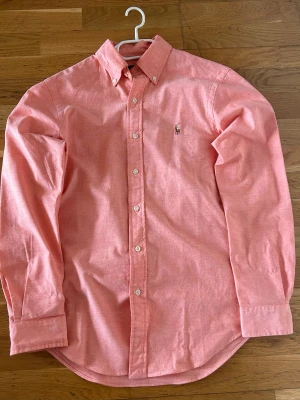 Korallfärgad skjorta från Polo Ralph Lauren - Säljer en snygg korallfärgad skjorta från Polo Ralph Lauren i slim fit stretch oxford. Skjortan har button-down krage, långa ärmar och den klassiska broderade loggan på bröstet. Perfekt för dig som gillar stilrena och bekväma plagg.