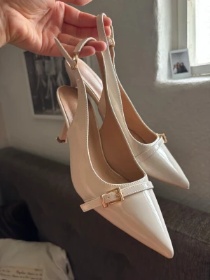 Vita lackade slingback-pumps - Säljer ett par vita pumps med lackad finish och spetsig tå. Skorna har en elegant rem med guldfärgat spänne över foten och öppen häl med justerbar slingback. Klacken är smal och medelhög, vilket ger en stilren siluett. Perfekta för att lyfta vilken outfit som helst.