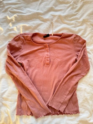 Rosa ribbad långärmad topp - Söt rosa långärmad topp med ribbad struktur och knappar framtill. Toppen har en lätt vågig kant nertill och vid ärmsluten, vilket ger en extra detalj. Perfekt att styla med jeans eller kjol för en avslappnad look.