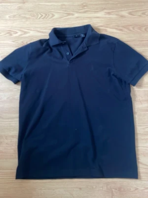 Svart pikétröja från BOSS - Klassisk svart pikétröja från BOSS med korta ärmar och krage med knappar. Diskret BOSS-brodyr på bröstet ger en stilren look. Perfekt till jeans eller chinos för en clean outfit.