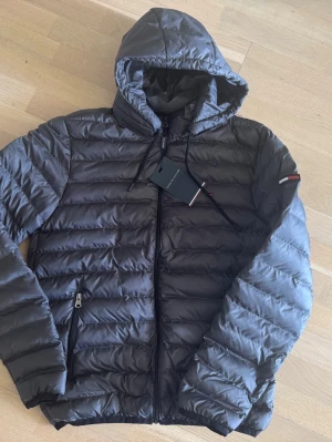 Mörkgrå Tommy Hilfiger dunjacka med avtagbar huva - Snygg mörkgrå pufferjacka med huva och dragkedja framtill. Jackan har två sidofickor med dragkedja och diskret detalj i rött, vitt och blått på ärmen. Fodret är delvis rött och materialet är glansigt och quiltat för extra värme.