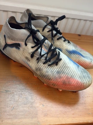  PUMA-FUTURE 8 PRO FG/AG - Snygga fotbollsskor från Puma med högt stickat skaft och svarta snören. Skorna har en unik färgkombination i vitt, blått och rött samt den klassiska Puma-loggan på sidan. Ovandelen är i syntetmaterial och ger en bra passform och kontroll på planen.  Skorna har synliga spår av användning och behöver rengöras, vilket syns på bilderna. Snörena är utbytta eftersom de gamla gick sönder. I övrigt är skorna fortfarande fullt användbara och passar perfekt för fotbollsträning och match.