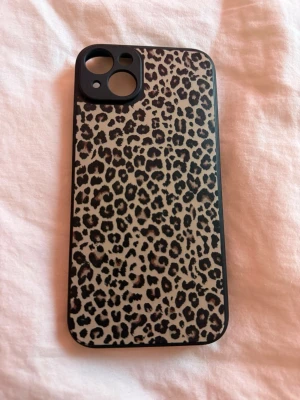 Mobilskal leopardmönster svart/brun  - Snyggt mobilskal med leopardmönster i svart och brun. Skalets kanter är svarta och det har öppningar för kamera och knappar. Mjuk plast likt silikon. iPhone 14 Plus 