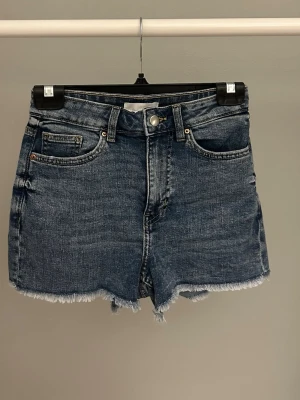 Blå högmidjade jeansshorts - Snygga blå jeansshorts med hög midja och råa, fransiga benslut. Klassisk femficksmodell med knapp och dragkedja framtill. Perfekta för varma dagar och ger en avslappnad vibe.