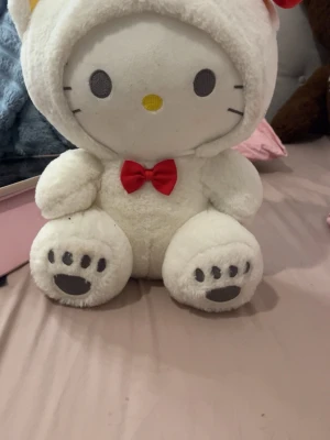 Stor vit Hello Kitty nalle - Söt och mjuk Hello Kitty gosedjur i vitt plyschmaterial med en röd rosett vid halsen och svarta tassavtryck på fötterna. Perfekt som dekoration eller mysig kompis i sängen.