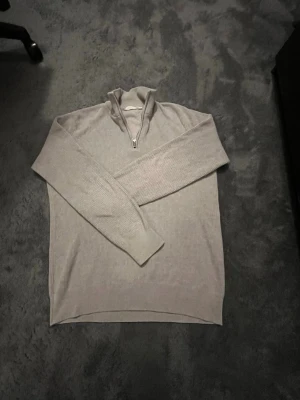  Ribbstickad tröja med half zip - Snygg grå/beige ribbstickad tröja med halv dragkedja och hög krage. Tröjan har långa ärmar och är tillverkad i ett mjukt, tunt material som passar perfekt för lager på lager. Enkel och stilren design som funkar till det mesta.