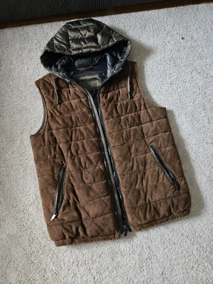Massimo Dutti Mocka Dunväst - Snygg Massimo Dutti brun dunväst med quiltat mönster och avtagbar huva i mörkgrått. Västen har två dragkedjeförsedda fickor framtill och stängs med dragkedja. Materialet är en äkta mocka och glansigt tyg på huvan och baksidan. Perfekt för lager på lager under kyliga dagar. Obs har en fläck på vänstra axeln se bild