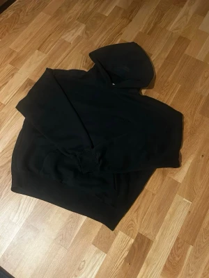 Svart Essentials Fear of God hoodie - Säljer en svart hoodie från Essentials Fear of God med stor huva och tryck på ryggen. Hoodien har en relaxed fit och ribbade muddar vid ärmslut och nederkant. Perfekt för dig som gillar streetwear och enkel stil. Passar som storlek M den är baggy också