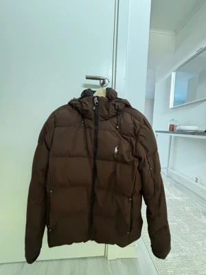 Ralph lauren jacka  - Snygg brun pufferjacka med huva och dragkedja framtill. Jackan har två sidofickor med dragkedja och en liten logga på bröstet. Perfekt för kalla vinterdagar och ger en riktigt trendig look.