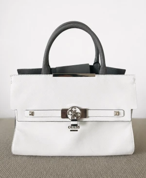 Birkin-inspirerad Guess Vit handväska med silverdetaljer - Nypris 2999kr - Stilren vit handväska från Guess med silverfärgat lås och detaljer. Väskan har två handtag i svart och vitt samt en dekorativ rem framtill. Klassisk rektangulär form och tillverkad i slätt konstläder.