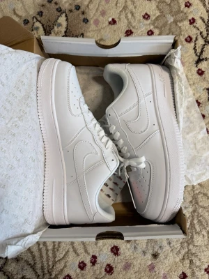 Nike Air Force 1 vita sneakers - Klassiska vita Nike Air Force 1 sneakers i lågt utförande. Skorna har en stilren design med perforerad tå, vita skosnören och den ikoniska swoosh-loggan på sidan. Tillverkade i slätt läder med en robust gummisula för extra komfort och streetstyle.