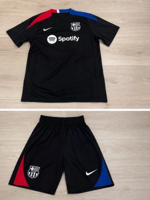 Svart FC Barcelona träningsset Nike - Säljer ett svart träningsset från Nike och FC Barcelona med klubbmärke och Spotify-logga på tröjan. Tröjan har röda och blå detaljer på axlarna, och shortsen har matchande färgblock på sidorna. Perfekt för fotbollsträning eller sportiga dagar.