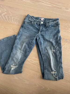 Blå slitna jeans med hål - Säljer ett par blå jeans från Gina tricot med slitningar och hål på benen. Jeansen har låg midja Materialet är denim i bomull och de har en avslappnad, trendig look.