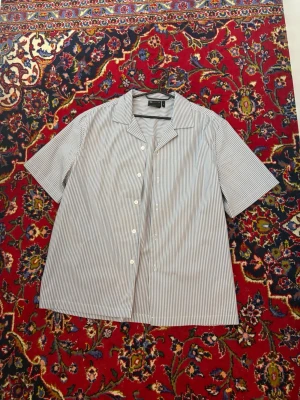 Randig skjorta från ASOS DESIGN - Kortärmad skjorta från ASOS DESIGN i vitt och blått med smala ränder. Skjortan har klassisk krage, knäppning framtill och en avslappnad passform. Perfekt för en stilren och enkel look.