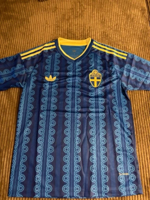 Blå Sverige fotbollströja Adidas - Säljer en blå fotbollströja från Adidas med Sveriges landslagsemblem på bröstet. Tröjan har gula detaljer, mönstrade ränder och klassiska Adidas-ränder på axlarna. Tillverkad i lätt och ventilerande material, perfekt för träning eller match.