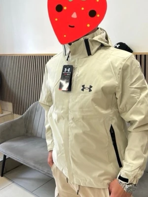 Beige vindjacka från Under Armour - Snygg beige vindjacka från Under Armour med svart logga på bröstet. Jackan har dragkedja framtill, två svarta dragkedjefickor och justerbar huva. Tillverkad i lätt och vattenavvisande material, perfekt för blåsiga dagar. Nu i förpackning 