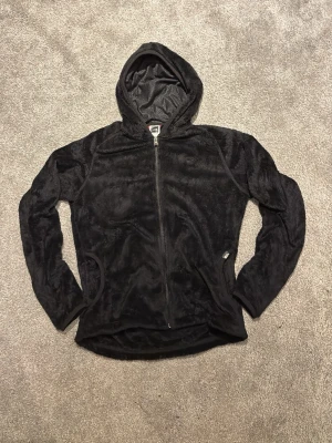 Svart North Face teddyjacka med huva - Mysig svart teddyjacka med dragkedja framtill och två fickor. Jackan har en skön huva och är gjord i mjukt, fluffigt material som håller dig varm. Perfekt för kyliga dagar när du vill vara både bekväm och snygg.