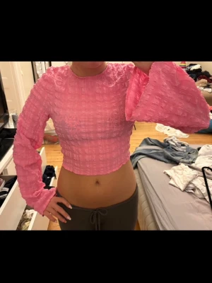 Rosa croppad topp med vida ärmar - Säljer en rosa croppad topp med långärmade vida ärmar, strukturerat tyg och öppen rygg. Toppen har en tight passform över kroppen med ett snöre längst med ryggen!!💗