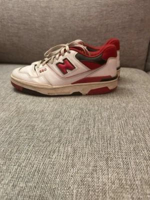 New Balance 550 vita och röda sneakers - Säljer ett par New Balance 550 sneakers i vitt och rött med svarta detaljer. Skorna har en klassisk låg siluett, snörning och NB-logga på sidan och hälen. Ovandelen är i läder och textil, sulan är vit med röda inslag. Perfekta för dig som gillar retrostil.