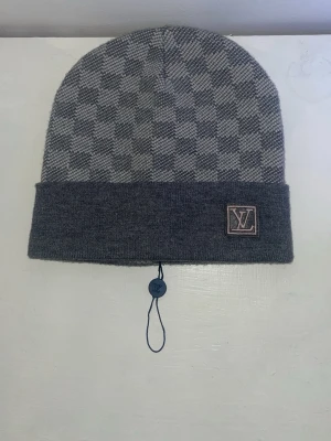 Grå vinter mössa  Damier Knit Beanie  - Snygg och exklusiv stickad mössa  i grått med det klassiska Damier-rutmönstret. Mössan har en vikt kant med -logotypen framtill och en diskret tagg kvar. Perfekt för höst och vinter för dig som vill ha både stil och värme.