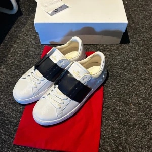 Vita sneakers med svart band - Snygga vita sneakers med ett brett svart band över snörningen och gulddetaljer på plösen. Skorna har en chunky vit sula och nitar på hälen för en edgy look. Tillverkade i slätt läder och har en modern, stilren design. Behöver andra snören för dem jag köpte till var inte bra…