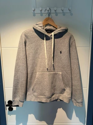 Ralph Lauren hoodie - Klassisk grå hoodie med luva och vita dragsnören. Har en broderad liten logga på bröstet och en stor känguruficka framtill. Tillverkad i mjukt sweatshirtmaterial som är perfekt för chill dagar. Ribbade muddar vid ärmslut och nederkant.