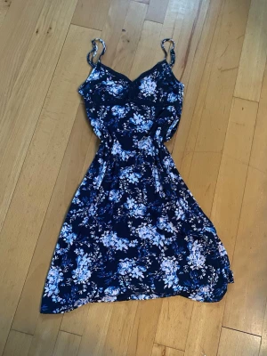 Blommigt nattlinne - This is a sleeveless, navy blue dress with a white and light blue floral pattern. It features thin spaghetti straps and a fitted bodice with a slightly flared skirt. The dress is laid flat on a wooden floor.