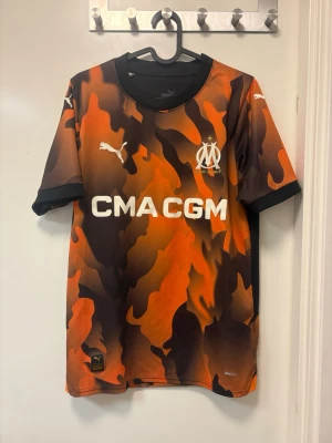 Marseille matchtröja orange camo Puma - Snygg Marseille matchtröja från Puma i orange, svart och brun camo-design. Tröjan har klubbmärke och Puma-logga på bröstet samt sponsortryck framtill. Tillverkad i ett lätt och ventilerande funktionsmaterial, perfekt för fotboll eller träning.
