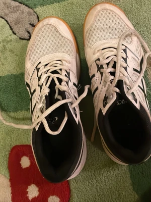 Asic inomhusskor - ett par Asics Upcourt 4 inomhusskor i vitt och svart med klassisk ljus gummisula (gumsole) anpassad för inomhussporter 