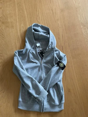 Grå zip hoodie från Stone Island - Snygg grå zip hoodie från Stone Island med klassisk logopatch på vänster ärm. Hoodien har dragsko i huvan, ribbade muddar och fickor framtill. Perfekt för en avslappnad och trendig stil. Materialet är mjukt och bekvämt bomullstyg.