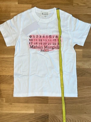 Maison Margiela vit t-shirt med tryck rosa - Maison Margiela t-shirt i vitt med ikoniskt siffertryck och rosa detalj framtill. Klassisk passform med korta ärmar och rund hals. Perfekt för dig som gillar streetstyle och vill sticka ut med ett coolt designerplagg.